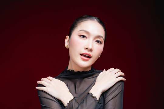 Dramatic close-up beauty portrait of a young Asian woman in a sheer black top gazing intensely at the camera against a deep red and black background.