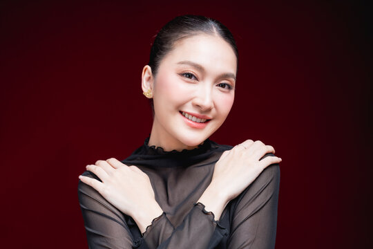 Dramatic close-up beauty portrait of a young Asian woman in a sheer black top gazing intensely at the camera against a deep red and black background.