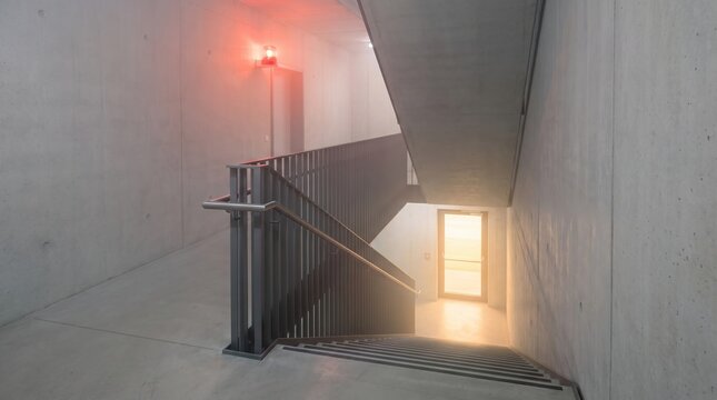 Modern industrial concrete emergency staircase interior with flashing red warning light and bright exit door for modern building fire safety and evacuation concept.