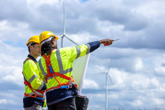 Multiethnic engineers holding blueprint and discussing green energy project. Wind turbine.farm worker pointing away while holding walkie-talkie with colleague. Professional industrial workers.