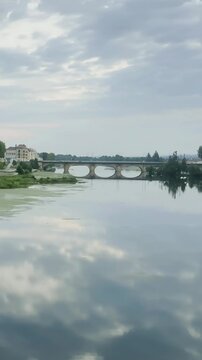 Calm river reflecting cloudy sky near stone bridge in town