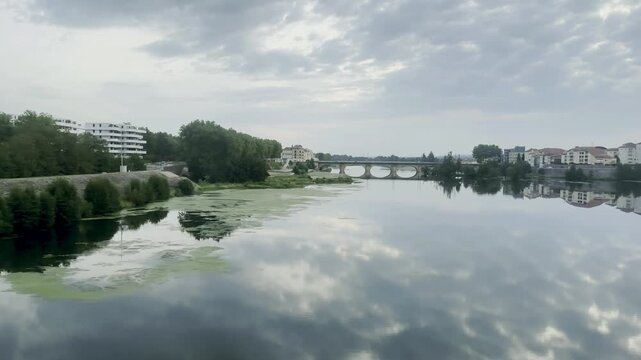 Calm river reflecting cloudy sky near stone bridge in town