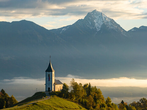 Aerial view of the Church of Saints Primus and Felician perched on a grassy hilltop surrounded by morning mist and dramatic mountains in Jamnik, Kranj, Slovenia.