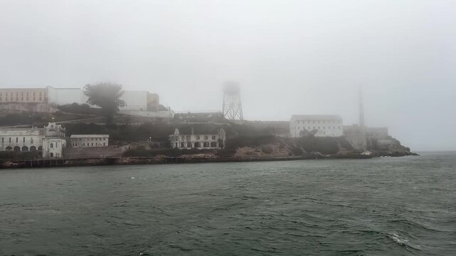 riding across San Francisco Bay, California, near the Golden Gate strait on a foggy day, while passing close by Alcatraz Island with buildings of famous United States Penitentiary, partly covered with