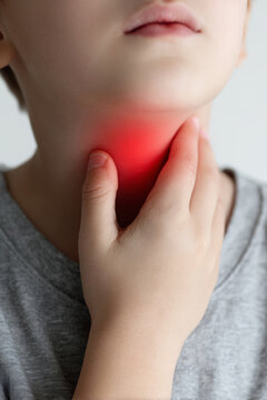 A child holds their throat with one hand in an indoor space. The child seems to be experiencing throat discomfort. The light is soft