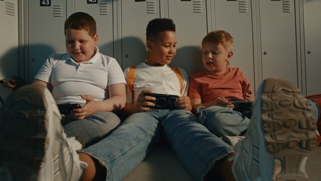 Elementary School Children Sitting in Sunny Corridor Beside Metal Lockers while Using Portable Gaming or Learning Devices. Modern Education, Digital Lifestyle, Childhood Communication. Handheld.