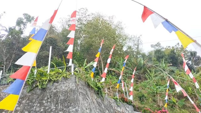 Colorful Indonesian red and white flags and decorative pennants waving along a rural roadside, celebrating Independence Day amid lush tropical vegetation and village scenery views. Umbul-umbul.