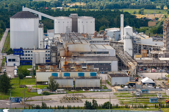 Aerial view of a large sugar plant processing sugar beets with industrial silos, cooling towers, and factory buildings surrounded by green fields and trees in Poland.