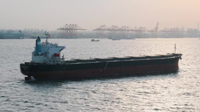 Bulk cargo vessel sailing through busy port waters with container terminals, cargo cranes and industrial facilities in Shanghai. Maritime transportation, international trade, logistics infrastructure