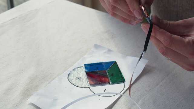 A male art student peels copper tape on top of a stained glass design during a small art class while sitting by a window.