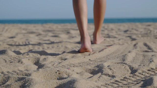 Closeup of foot of woman walking on sand during beach holiday, barefoot female legs stepping on warm golden shoreline toward beautiful blue ocean water under bright summer sun.
