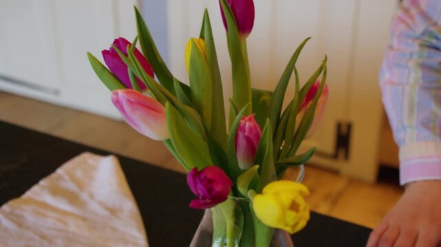 Woman pushes forward a clear vase with a finished floral arrangement containing pink, yellow, and red flowers for spring decoration.