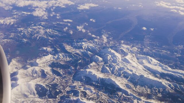 View from an airplane window capturing a vast snow covered mountain range with clouds drifting above rugged peaks.