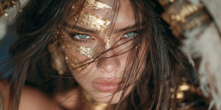 beautiful female warrior with captivating gaze and textured gold leaf face paint, young woman portrait with tousled dark brown hair, powerful fantasy warrior portrait featuring gold leaf makeup