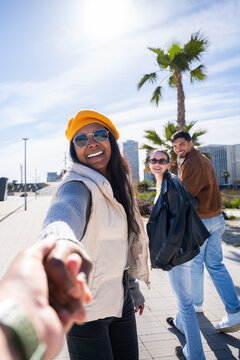Vertical. Happy Black woman smiles and reaches forward to lead the viewer by hand, while friends stand behind on a sunny promenade in a modern urban area.