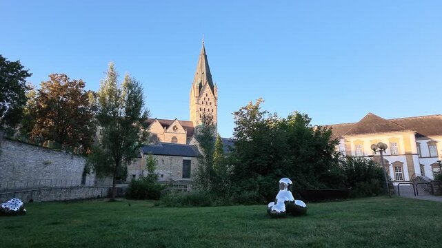 Right to left side tracking view crosses the cathedral meadow, reflective art structures, Paderborn Cathedral and the historic City Library.