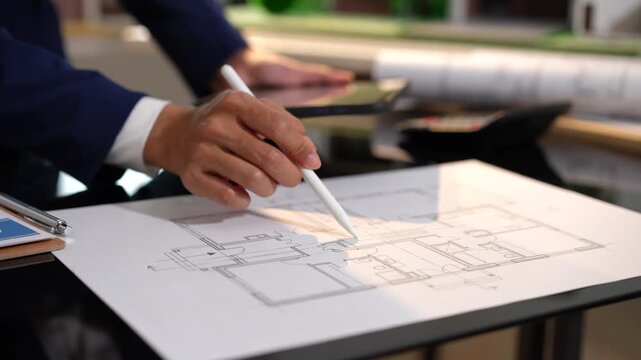 Close-up of a professional reviewing a house floor plan and using a digital pen on paper with a tablet nearby, showing planning, design, and real estate work in a modern office.