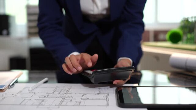 Architect or business professional using a calculator over building plans on a desk, with a tablet nearby in a modern office, showing budgeting, planning and project work.