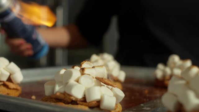 Marshmallow topped cookies are toasted with a blue culinary torch flame on a baking tray