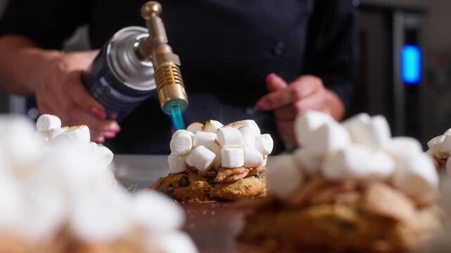 Culinary torch browns white marshmallows piled over cookies on a metal baking surface