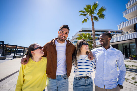 Multicultural friends stand together and laugh in a bright residential area with palm tree, blue sky and contemporary apartment buildings.