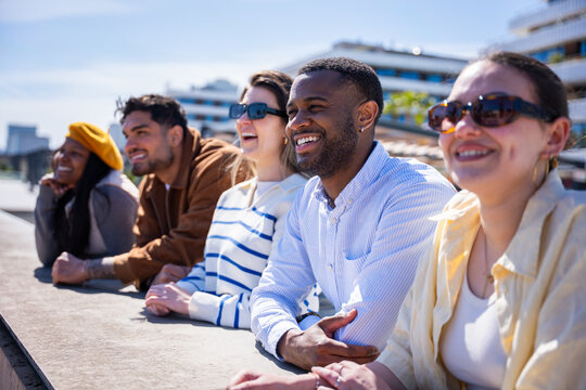 Group of multicultural friends enjoy a bright sunny day while leaning on a ledge in a contemporary residential neighborhood with modern architecture.