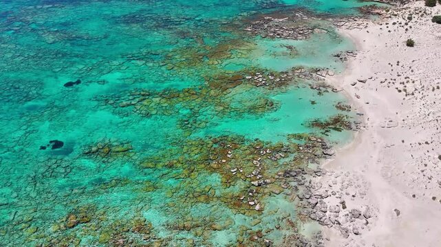 The aerial camera tracks Elafonissi reef beside a pale sandy shoreline, revealing clear turquoise shallows, submerged rock patterns and scattered beach visitors.