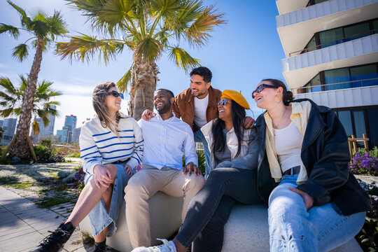 Diverse friends sit close together in a bright neighborhood and smile during a relaxed outdoor social moment with palm trees and modern architecture.