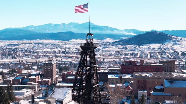 Drone footage above Butte, Montana, framing a memorial structure with an American flag waving against the backdrop of a snow-covered winter cityscape and surrounding rolling hills