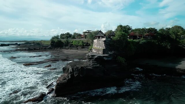 Cinematic aerial shot of Tanah Lot Temple in Bali, Indonesia, captured at high tide, highlighting the temple's precarious position on a rock formation against the crashing ocean waves fvp drone