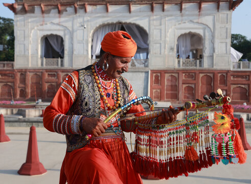 Lahore, Punjab, Pakistan - 11 April 2025: View of a traditional musician in vibrant orange attire performing with a decorated string instrument at the historic Shalamar Garden.