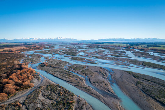 New Zealand - 09 July 2021: Aerial view of a braided river flowing through a vast landscape with snow-capped mountains in the background under a clear blue sky.