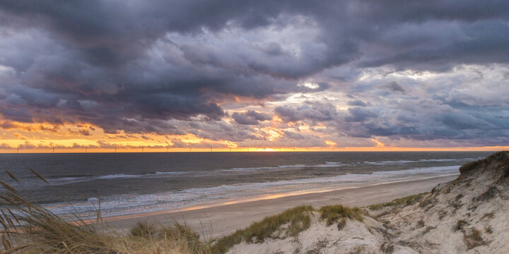 Aerial view of the sandy coastline with dunes, marram grass, and a dramatic sunset over the North Sea with distant wind turbines on the horizon in Hvide Sande, Denmark.