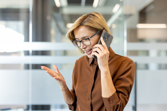 Woman in glasses talking on her smartphone, expressing anger and stress during a difficult conversation, gesticulating with her hand while standing in a modern office environment