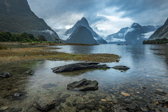 New Zealand - 24 April 2022: View of Mitre Peak rising above the calm waters of Milford Sound with rocky shores and dramatic clouds.