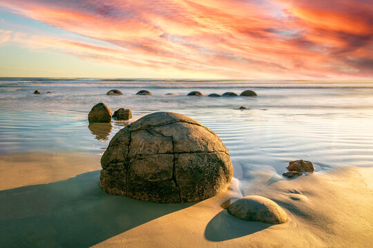 New Zealand - 07 December 2020: View of the Moeraki Boulders on the sandy beach with waves and a vibrant sunset sky.