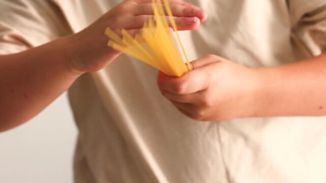 Little boy hands twirl a bundle of dry uncooked spaghetti noodles, spinning the raw strands playfully as part of imaginative pretend cooking play in a warm, softly lit, cozy kitchen setting at home.