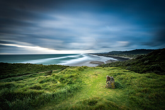 New Zealand - 10 October 2021: View of a long exposure coastal landscape with rolling green hills in the foreground and a vast ocean under a dramatic cloudy sky in New Zealand.