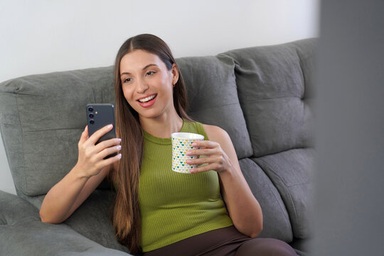 Happy young woman relaxing on couch with smartphone and cup of tea, mindful digital detox at home