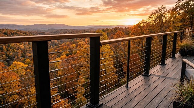 Deck railing of black metal posts and steel cables with wooden handrail, overlooking autumn forest of golden orange foliage at sunset