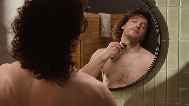 Young Caucasian man shaving neck with electric trimmer in bathroom mirror, rear view. Clip is suitable for male grooming product commercials and skincare brand advertisements