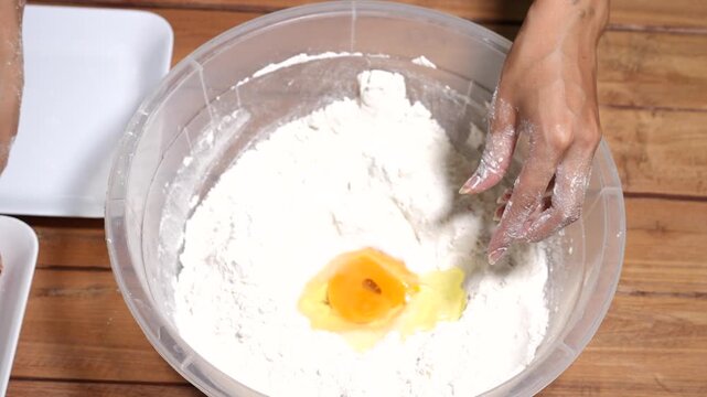 Close-up of hands mixing flour and egg in a bowl on a wooden table, showing homemade dough preparation and simple home baking in a warm kitchen setting.