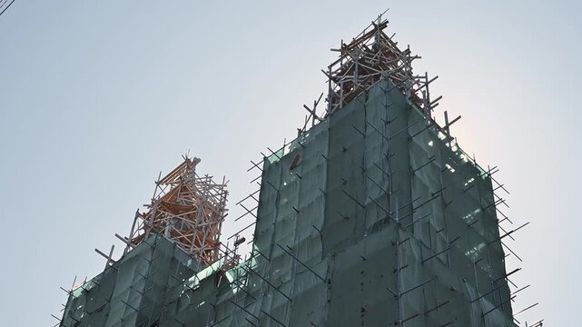 constructing a tall building structure surrounded by scaffolding and cranes. The activity occurs under clear skies during daytime, showcasing the construction process