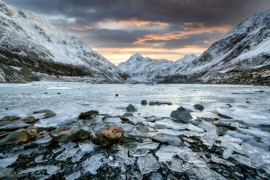 New Zealand - 10 July 2021: View of Aoraki Mount Cook with ice fragments on the shore of a glacial lake under a dramatic sunset sky.