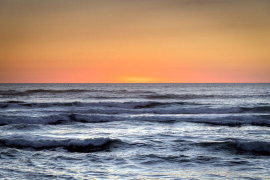 New Zealand - 13 March 2021: View of ocean waves rolling towards the shore under a vibrant orange sunset sky.