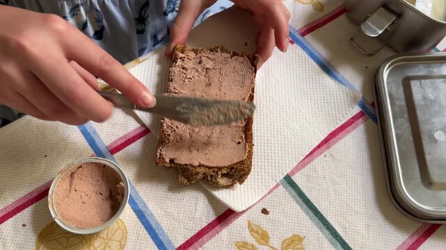 Horizontal top-down close-up shot showing the detail of a girl's hands spreading pate on a thick slice of bread with a knife.