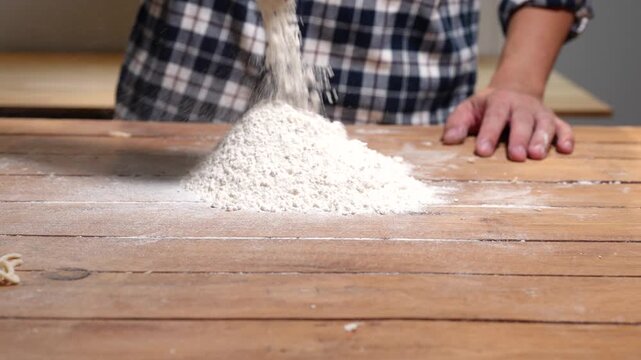Close view of flour falling onto a wooden table with a baker in a plaid shirt, showing homemade baking, cooking preparation, and rustic kitchen work.