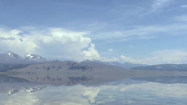 Witness Khuvsgul Lake's pristine waters in Mongolia, perfectly reflecting towering mountains and expansive clouds. A breathtaking tableau of nature's artistry unfolds under a bright, clear daytime sky