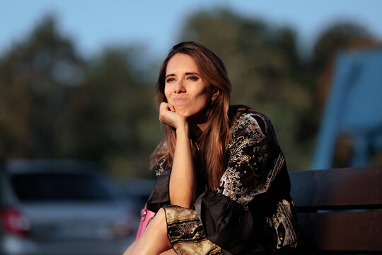 Thoughtful Woman Sitting On Bench Resting Chin On Her Hand. Pensive lady sitting outdoors with a dreaming expression looking at camera