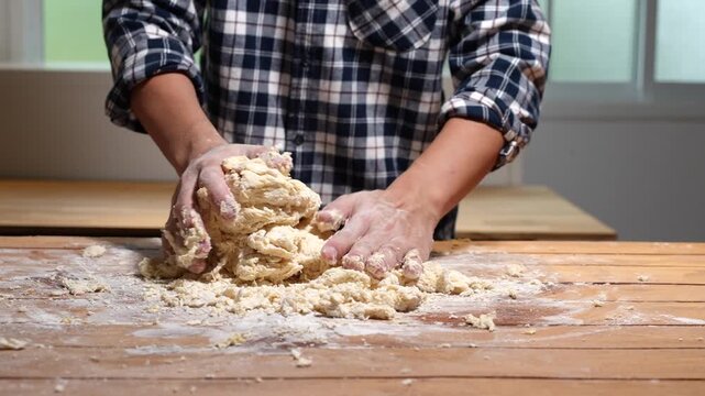 Close view of a person kneading fresh dough on a wooden table in a home kitchen, with flour scattered around, showing handmade baking, cooking and rustic food preparation.